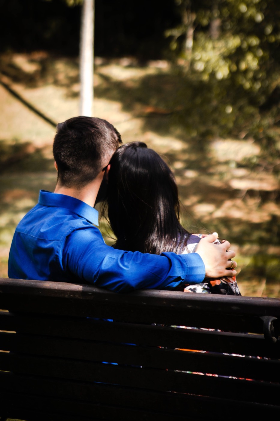 A couple sits close together on a park bench facing away toward trees