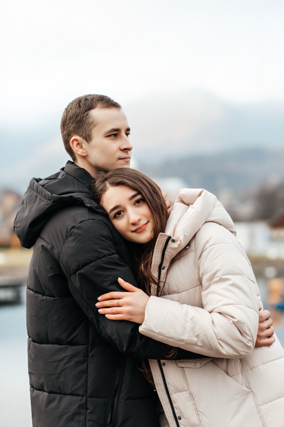 A couple in winter coats embrace outdoors with soft daylight on their faces