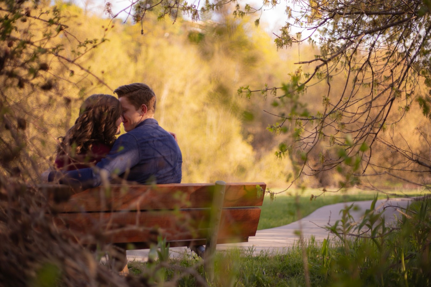 A couple sits close on a wooden park bench with their foreheads touching under warm sunset light
