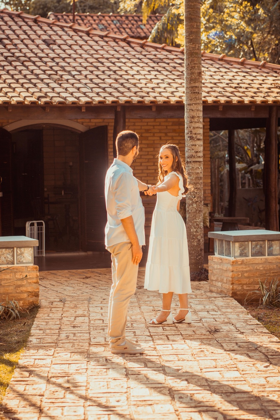 A smiling woman in a white dress looks back while holding hands with a man on a sunlit stone path
