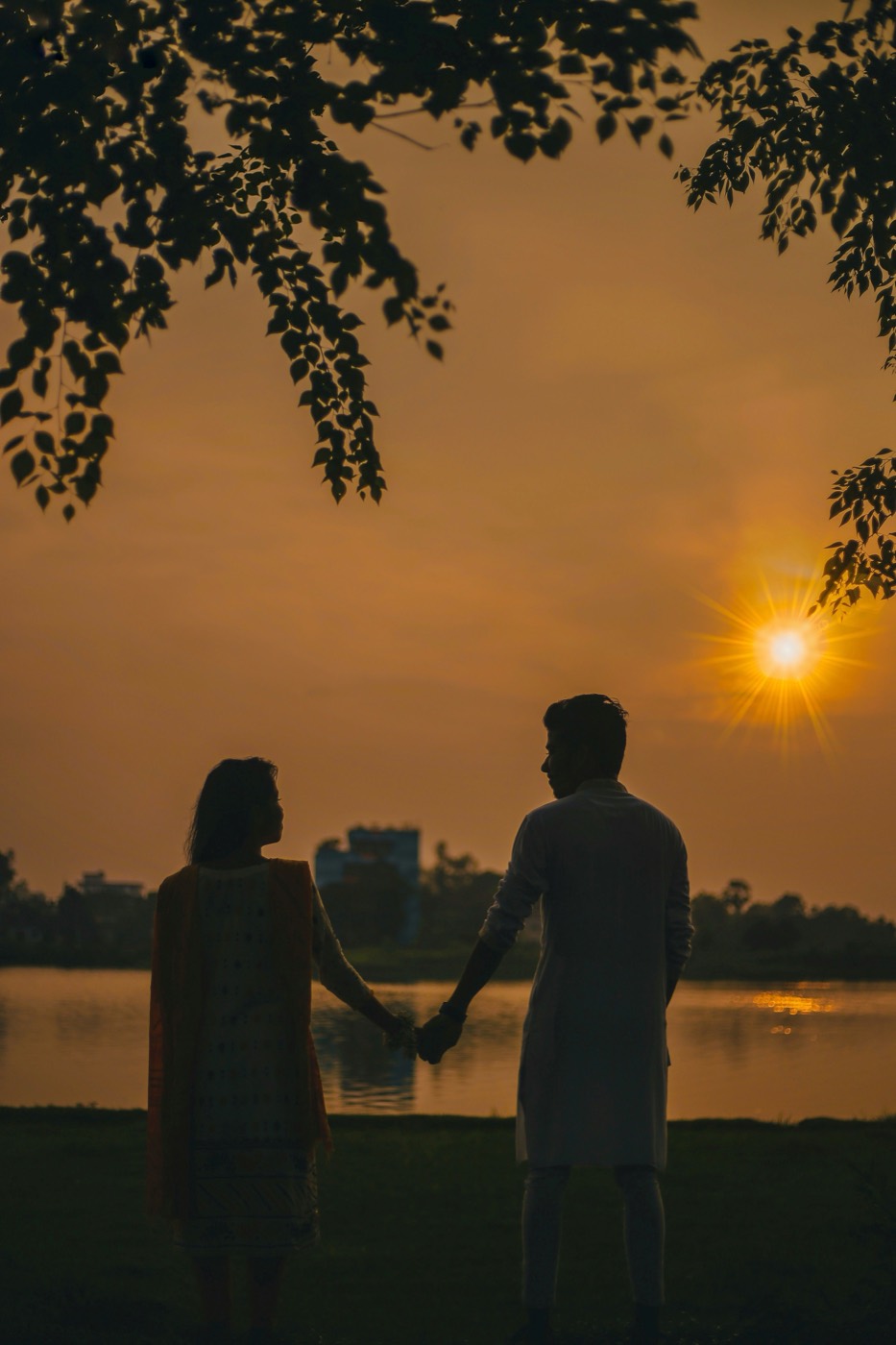 A couple holds hands beside a lake at sunset with warm light on the water