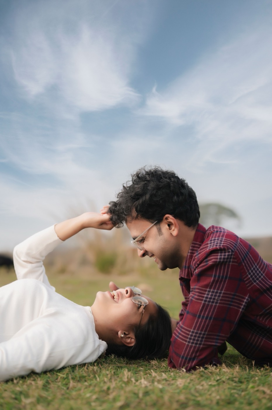 A man in a plaid shirt leans over a laughing woman in glasses as they lie together in a grassy field