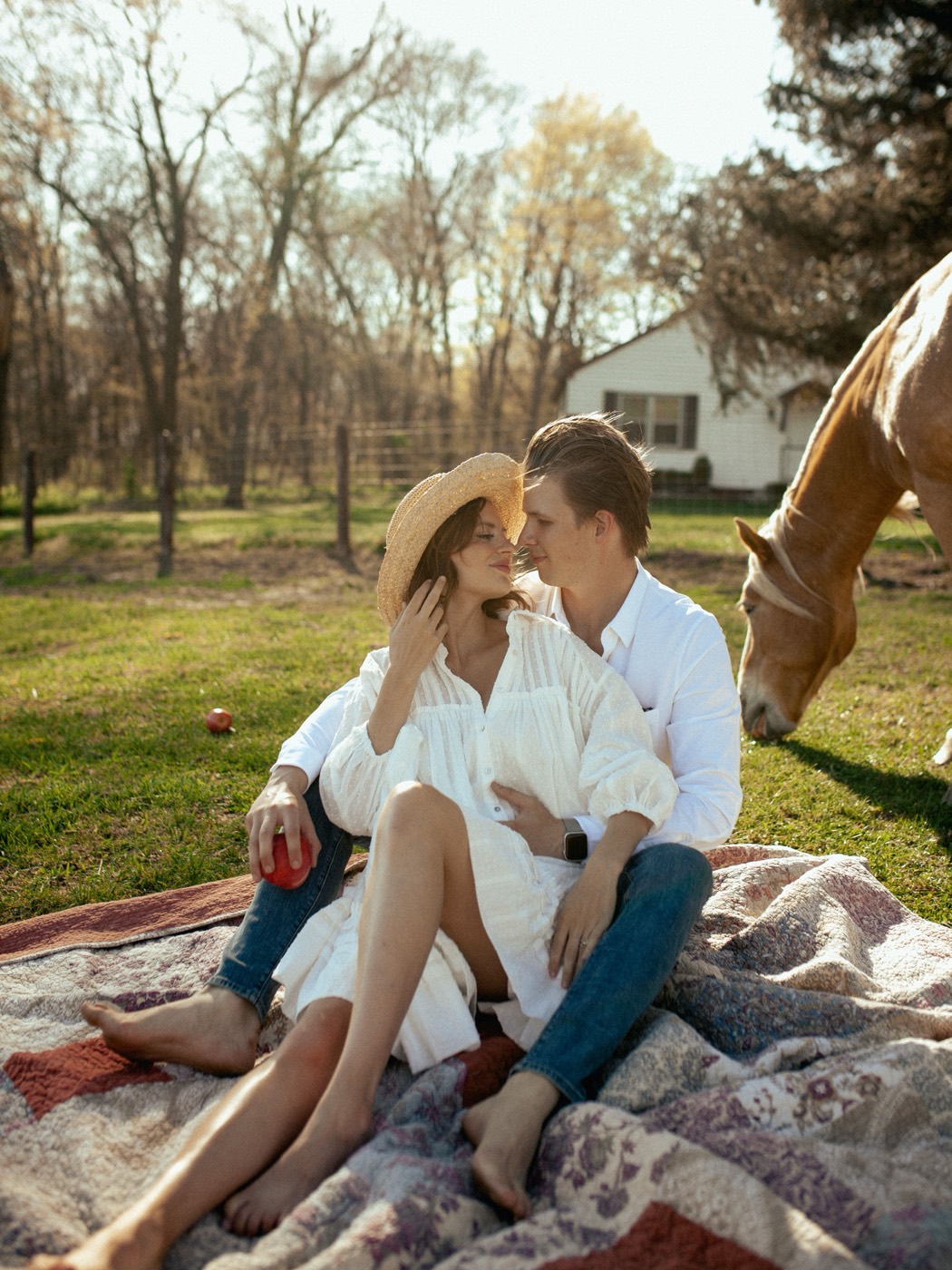A couple sits on a picnic blanket near a horse in a sunlit farm field