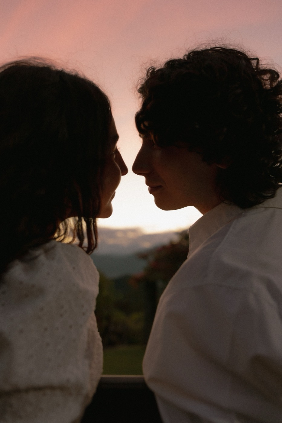 A man and woman stand in profile at a dusk overlook with soft sky behind them