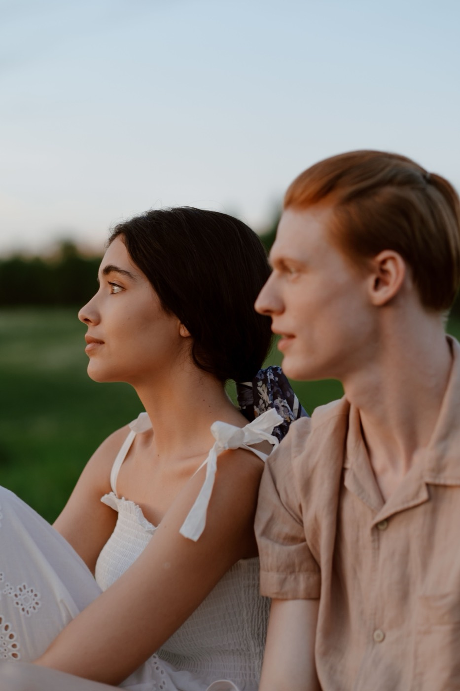 A couple sits close together in a grassy field at twilight with soft sky light behind them