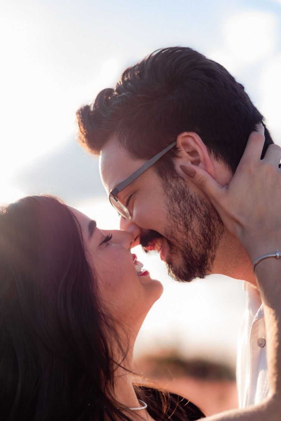 A smiling man with glasses and a woman with long dark hair touch foreheads in warm golden-hour light