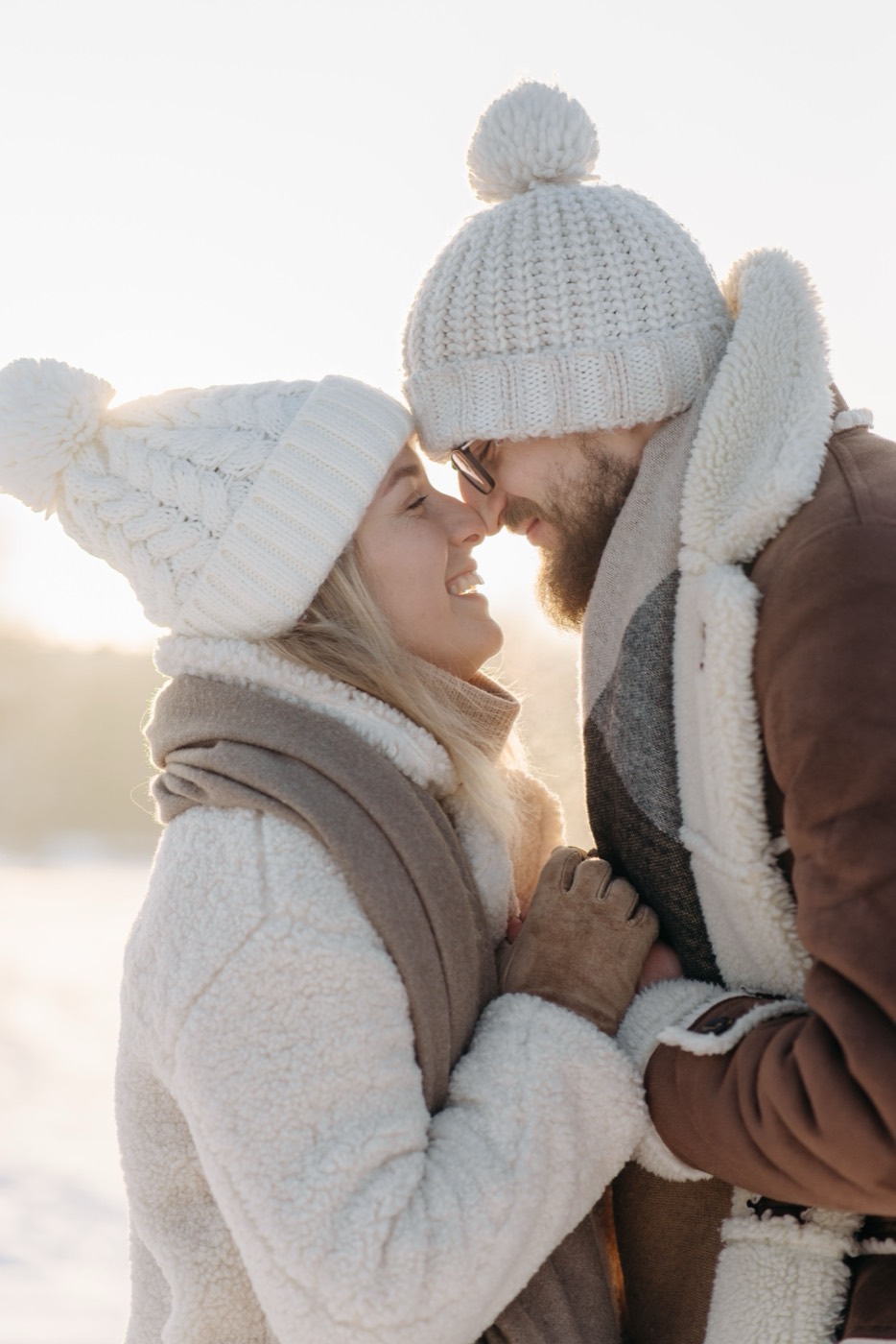 A bearded man in glasses and a blonde woman in white knit hats lean their foreheads together in the snow